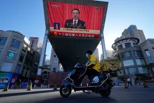 Un repartidor pasa en moto por delante de la pantalla de un centro comercial que emite en directo el discurso del primer ministro de China, Li Keqiang, en la sesión inaugural de la Asamblea Popular Nacional de China, en Beijing, el 5 de marzo de 2022. (AP Foto/Ng Han Guan)