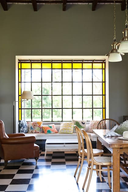 Igual que en living, el ventanal del comedor tiene vidrio repartido con guarda amarilla que suaviza la entrada de luz. Mesa con base de cobre y tapa de mármol y una taza gigantesca que se usa como florero.