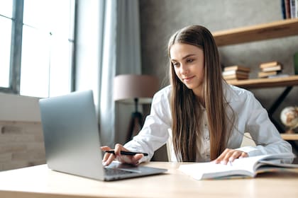 A beautiful caucasian female student is studying in college remotely. She is sitting at a table at home with a laptop and a notepad and concentrated is watching a video conference lesson