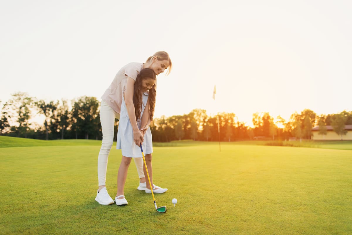 A woman is teaching her daughter to play golf. They are standing on the golf course, the girl is holding a golf club, the woman is standing behind her and directs her to strike