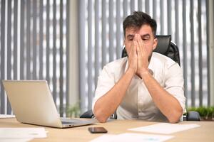 A young leader sits at his workplace in the office and looks tiredly at one point, hiding his face in his hands. Manager tired of work