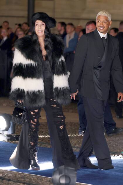 Acompañada por su novio Alexander Edwards, Cher se robó todas las miradas con sus jeans acampanados con roturas que complementó con maxiabrigo de pelo bicolor y gorra.