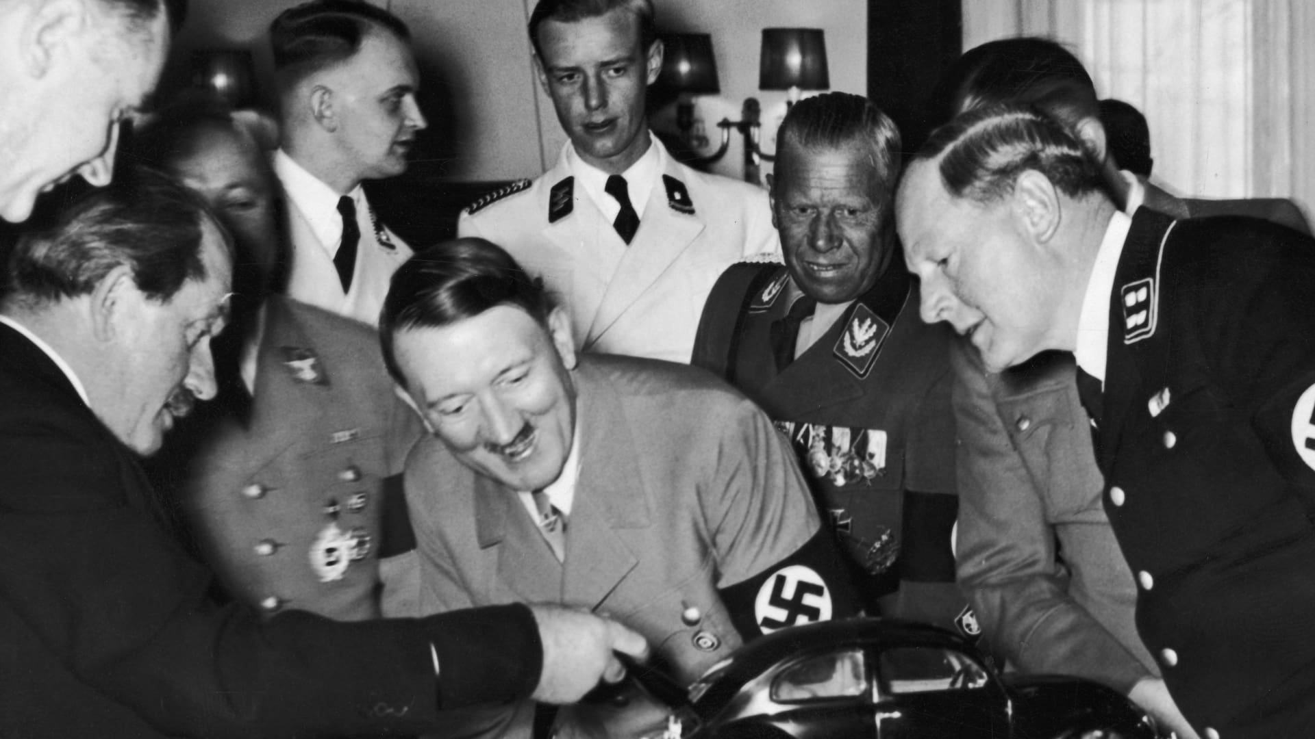 Adolf Hitler observa una maqueta del "auto del pueblo" y se ríe al descubrir al motor en la parte trasera. Quien levanta la tapa del baúl es el diseñador Ferdinand Porsche