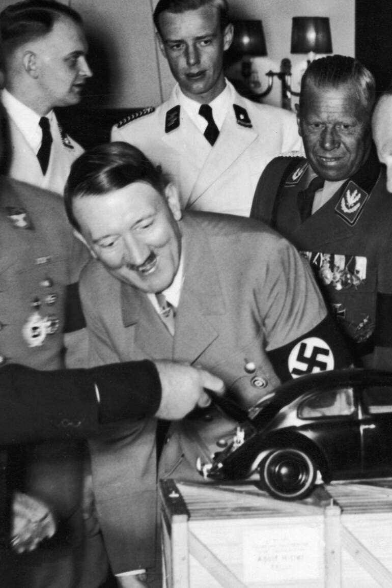 Adolf Hitler observa una maqueta del "auto del pueblo" y se ríe al descubrir al motor en la parte trasera. Quien levanta la tapa del baúl es el diseñador Ferdinand Porsche