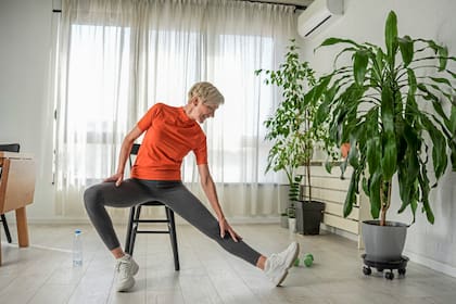An elderly woman in an orange shirt and gray leggings sits on a chair, stretching her leg to the side. She reaches toward her foot in a bright living room with plants, promoting senior wellness