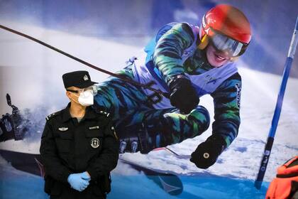 ARCHIVO - Un agente de policía con mascarilla y gafas protectoras contra el COVID-19, de pie ante un cartel con la imagen de una esquiadora en una estación de tren en Zhangjiakou, en la provincia norteña china de Hebei, el viernes 26 de noviembre de 2021. (AP Foto/Mark Schiefelbein, Archivo)