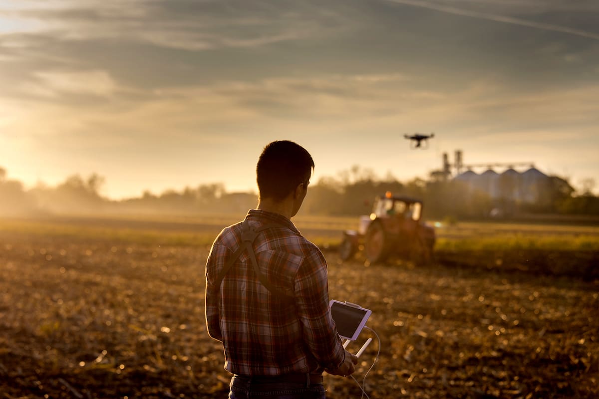 Attractive farmer navigating drone above farmland with silos and tractor in background. High technology innovations for increasing productivity in agriculture