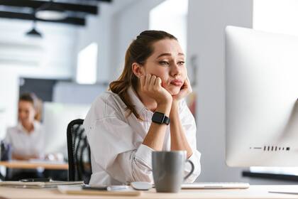 Bored young woman dressed in shirt sitting at her workplace at the office, looking at computer