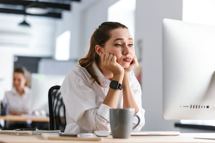Bored young woman dressed in shirt sitting at her workplace at the office, looking at computer