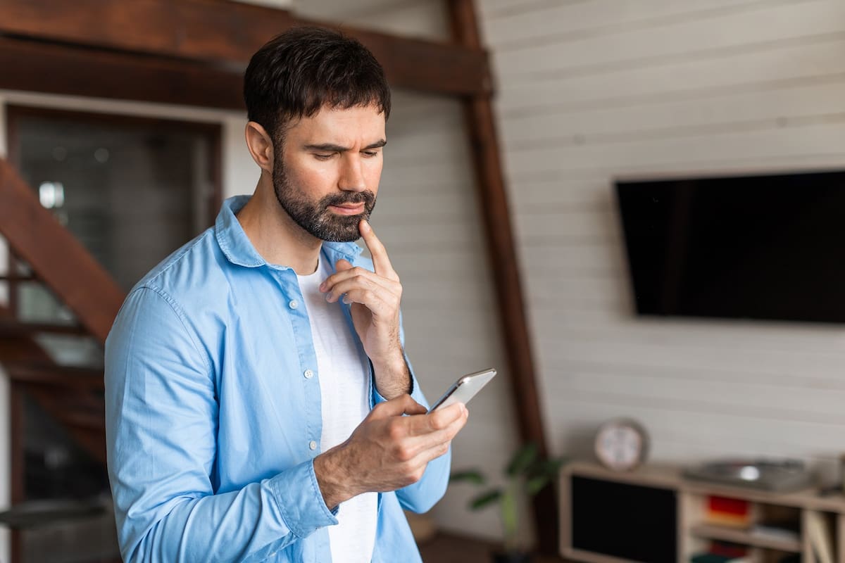 Confused man in a blue shirt is focused on his cell phone screen, appearing to read or type a message. He is standing in a casual posture, possibly outdoors.