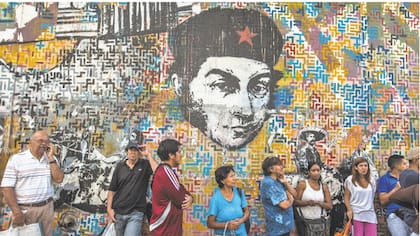 Consumidores hacen fila para comprar alimentos en Caracas bajo la mirada de un Simón Bolívar con boina.