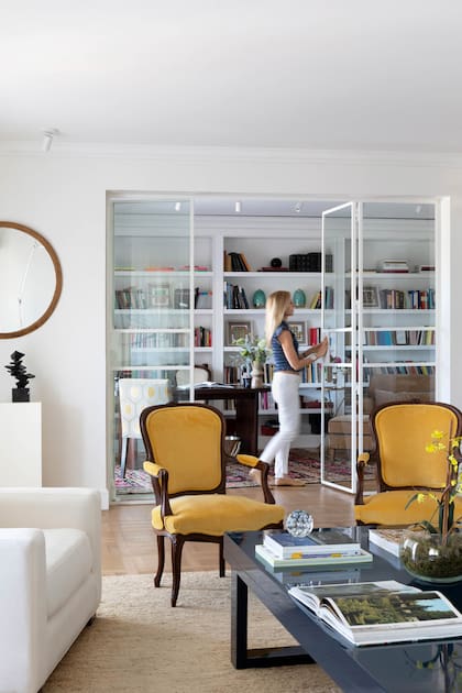 Detrás de los sillones tapizados en pana amarilla, un cerramiento de vidrio plegable integra el escritorio, con mesa de madera maciza.