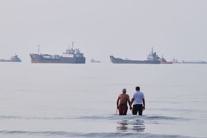 Dos hombres caminan en las aguas del estrecho de Ormuz con barcos fondeados más lejos, cerca de Bandar Abbas, Irán