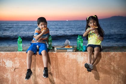 Dos niños comen pizza en el Malecón de Puerto Vallarta; en el cielo, los colores del atardecer