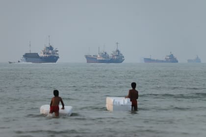 Dos niños de pie en aguas poco profundas con flotadores de espuma, mientras se ven buques de carga y otras embarcaciones en el estrecho de Ormuz, frente a Bandar Abbas, Irán, el martes 30 de junio de 2026. (Amirhosein Khorgooi/ISNA vía AP)
