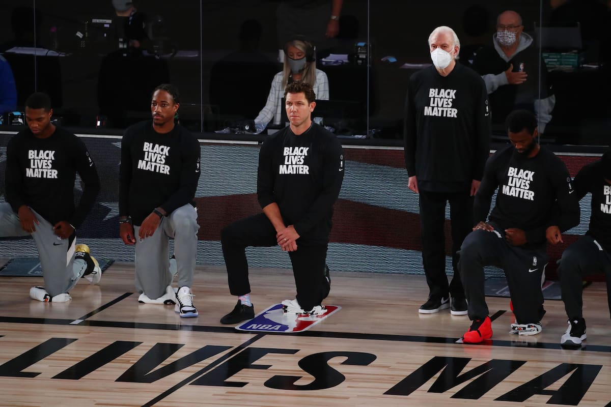 El entrenador en jefe de los San Antonio Spurs, Gregg Popovich, se pone de pie con una máscara mientras que el entrenador en jefe de los Sacramento Kings, Luke Walton (centro), se arrodilla con los jugadores antes de un partido de baloncesto de la NBA en el Visa Athletic Center en el ESPN Wide World