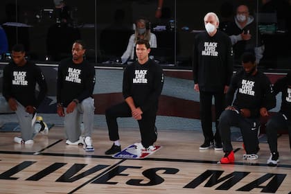 Gregg Popovich, DT de los Spurs, se mantiene en pie mientras que el entrenador en jefe de los Sacramento Kings, Luke Walton (centro), se arrodilla con los jugadores antes del partido de anoche en la NBA