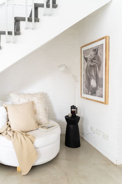 El espacio de abajo de la escalera muta: primero tuvo la pared de ladrillo a la vista, con un sillón gris con respaldo de capitonné; ahora, todo pintado de blanco, hay una poltrona circular con una lámpara y una mesa de apoyo (Gloria Atelier Deco).