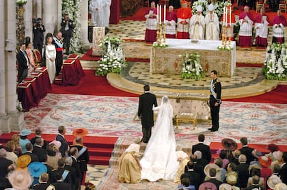 El ingreso de la novia a la catedral de la Almudena con el espectacular vestido obra de Manuel Pertegaz (1918-2014). Cuello en uve de resaltos volados o ‘corola’, en hilo de plata y oro platinado. Bordado con motivos como la flor de lis (versionada en vegetal y heráldica), espigas de trigo y madroños y tréboles. Las mangas, largas, estrechas y terminadas en sobre puños o manoplas con quilla y con los mismos bordados. Por delante, un triángulo subía desde el ruedo con los motivos dispuestos en rampante, y en la cola, lo mismo pero abriéndose proporcionalmente. El corte se prolongaba desde los hombros, facilitando el entalle hasta llegar a la cintura, desarrollándose radialmente hasta la envergadura de la cola: 4,5 metros a partir de la cadera, con un perímetro de 16 metros. Una obra de arte.