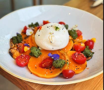 El pesto casero, las hierbas aromáticas, semillas de sésamo y flores comestibles seducen en esta burrata de Merienda