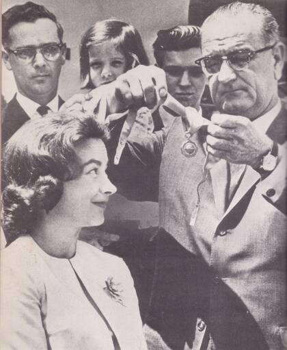 El presidente Lyndon B. Johnson condecora a Jerrie Mock en el Rose Garden de la Casa Blanca, el 4 de mayo de 1964. Detrás de la aviadora, observan la escena con mucha emoción su marido, Russell Mock, y sus hijos, Valerie y Gary. La medalla de oro, otorgada por la Agencia Federal de Aviación, es la máxima condecoración para un aviador civil en los Estados Unidos.