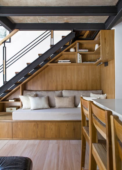 El sector de bajo escalera fue diseñado a medida, con un revestimiento en madera que contiene sillón, cajonera y biblioteca. Los almohadones y fundas se pensaron siguiendo la paleta neutra de todo el departamento.