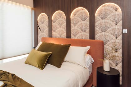 En el dormitorio del espacio de Halesa, la pared revestida con melamina Merlot (Faplac para Casa Leiro) tiene arcos calados retroiluminados que dejan ver un empapelado con motivos de abanicos (Tintachini, aplicado por Empapelarte). Cabecera de cama en terciopelo terracota y almohadones en tonos oliva.