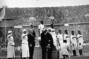 En el escalón más alto, Andrés Gazzotti, el capitán argentino, luce su corona de hojas de roble como campeón olímpico de Berlín 1936; más atrás, Roberto Cavanagh y jugadores de Gran Bretaña y México, mientras integrantes de la Agrupación Damas de Honor realizan el saludo nazi en la ceremonia de premiación en el Olympiastadion.