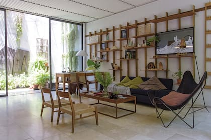 En el living, sofá oscuro, lámpara de pie con pantalla cobre, sillón BKF de piel, sillas bajas ‘Museo’ y mesa de lapacho ‘Bandeja’ (todo de Net Muebles) con ramas verdes en jarrón de cristal (Otra Sudestada).