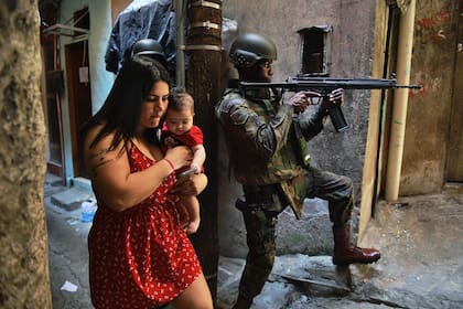 En esta foto de archivo tomada el 23 de septiembre de 2017, una mujer camina con su bebé frente a un soldado de la policía militarizado en posición y apuntando con su rifle en la favela Rocinha en Río de Janeiro, Brasil