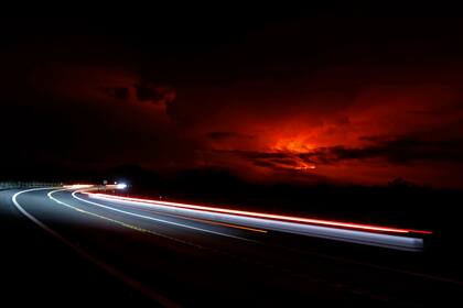 En esta fotografía de exposición prolongada, los automóviles avanzan por Saddle Road mientras el volcán Mauna Loa hace erupción a lo lejos, el lunes 28 de noviembre de 2022, cerca de Hilo, Hawai. (AP Foto/Marco Garcia)