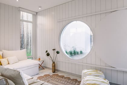 En esta sala de estar, la ventana circular (que da al patio del frente) se puede cerrar con un panel corredizo de madera pintada de blanco. Muro revestido de placa de fibrocemento pintada de blanco.
