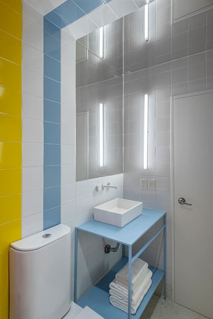 En este baño, se apostó por una estética lúdica, que eventualmente podría funcionar en un baño infantil. Las guardas de azulejos dibujan arcos que dan la vuelta entera. Se eligió apoyar la bacha sobre un soporte de hierro celeste.