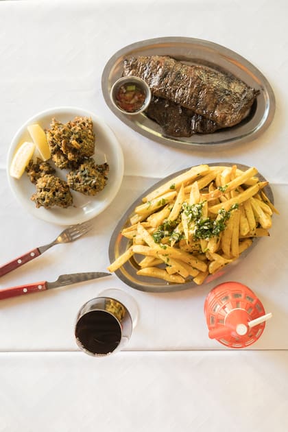 Entraña con papas fritas a la provenzal, buñuelos, vino y sifón en la mesa.