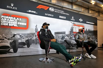 La imagen enlaza el pasado y el presente de la Fórmula 1: Lewis Hamilton y Valtteri Bottas, durante una conferencia de prensa del Gran Premio de Eifel; atrás, los autos aceleran en el viejo circuito de Nürburgring.