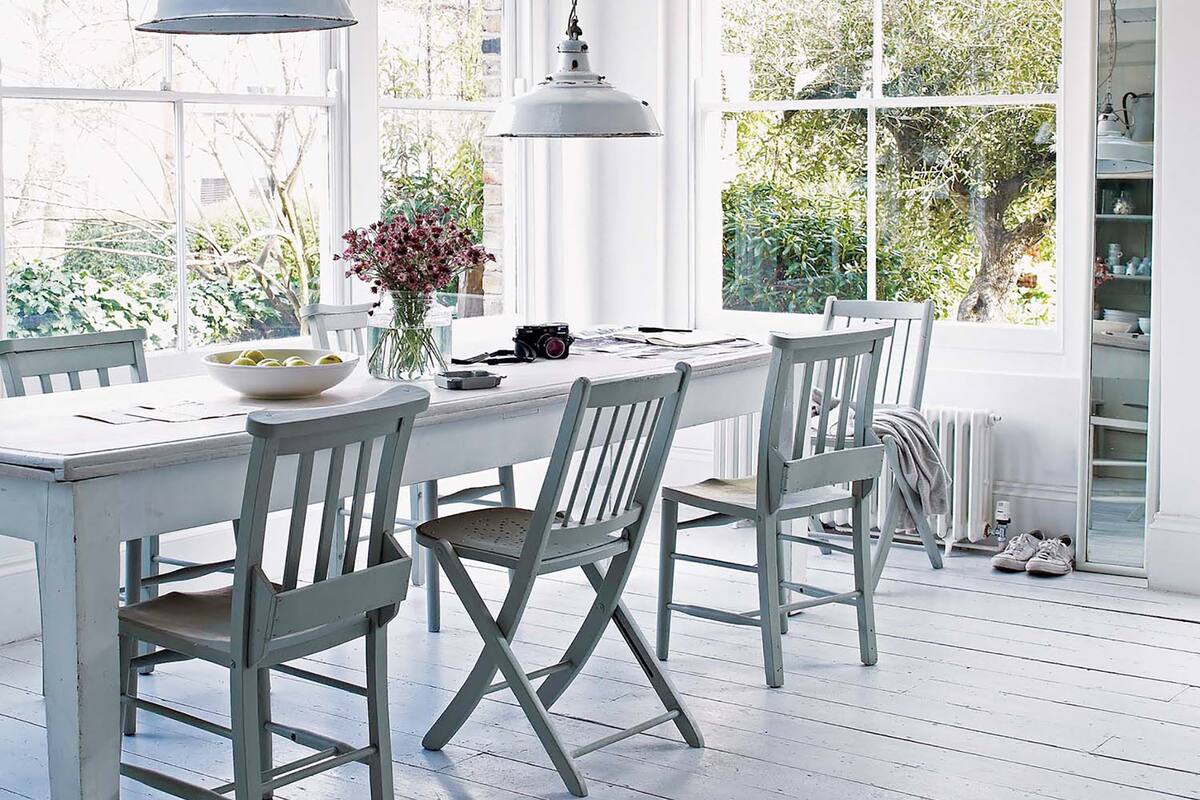 Foto de un comedor con paredes vidriada que dan al jardín, mesa y sillas de madera pintadas de blanca y lámparas colgantes.