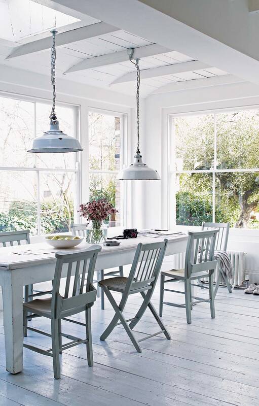 Foto de un comedor con paredes vidriada que dan al jardín, mesa y sillas de madera pintadas de blanca y lámparas colgantes.