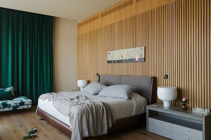 En la pared de la cama, revestimiento varillado. Respaldo color visón. Obra ‘Los arcanos’ de Paola Vergottini. Cortinas de pana. Sillón tapizado con géneros de Ronchamp. Mesas de luz (Matriz Design). Lámparas ‘Behaven’ (Foscarini).