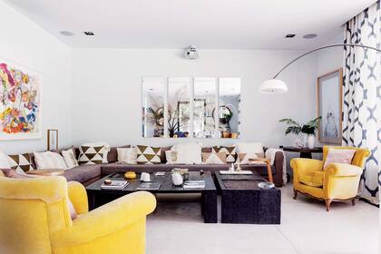 Sofá en ‘L’ tapizado con género en tonos uva (Agustina Lanusse) con almohadones de estampas geométricas (Didot), Sillones de corte retro (Jazmeen Deco) tapizados en terciopelo color mango (Agustina Lanusse). Mesas bajas negras (Magdalena y Bernarda Álvarez). Cuarteto de espejos (Harturo). Obra de arte de Zelmira Peralta Ramos. Cortinas estampadas (Didot).