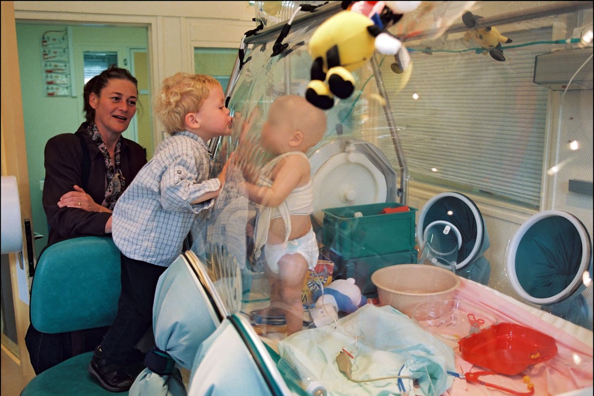 FRANCE - APRIL 28: Cured Wilco gives a kiss through the plastic sheet of the sterile 'bubble' to Yacine, another child affected with severe combined immunodeficiency (SCID) X1 who got a classical bone marrow transplant at Necker Hospital for Sick Children, in Paris, France on April 28, 2002, where Wilco got his first gene therapy in October 1999 - On the left is Roelien Conradi, Wilco's mother. (Photo by Raphael GAILLARDE/Gamma-Rapho via Getty Images)