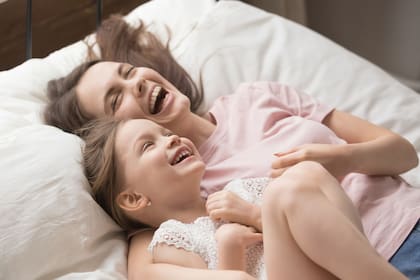 Funny overjoyed sisters having fun on weekend lying in bed in the morning on soft pillows laughing feels happy, mother and daughter enjoying time together. Leisure activities indoors with kids concept