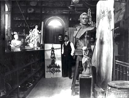 Gabriel Simonnet Dubois enseña una fotografía de la Araña del Centenario, con probabilidad en el taller de Azaretto. La maqueta del mismo artefacto se observa colgada detrás del busto de San Martín. Circa 1913.