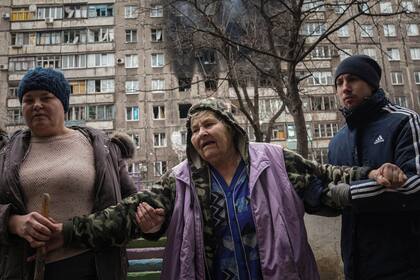 Gente ayudando a una anciana a caminar por la calle junto a un edificio de apartamentos alcanzado por proyectiles en Mariúpol, Ucrania, el lunes 7 de marzo de 2022. (AP Foto/Evgeniy Maloletka)