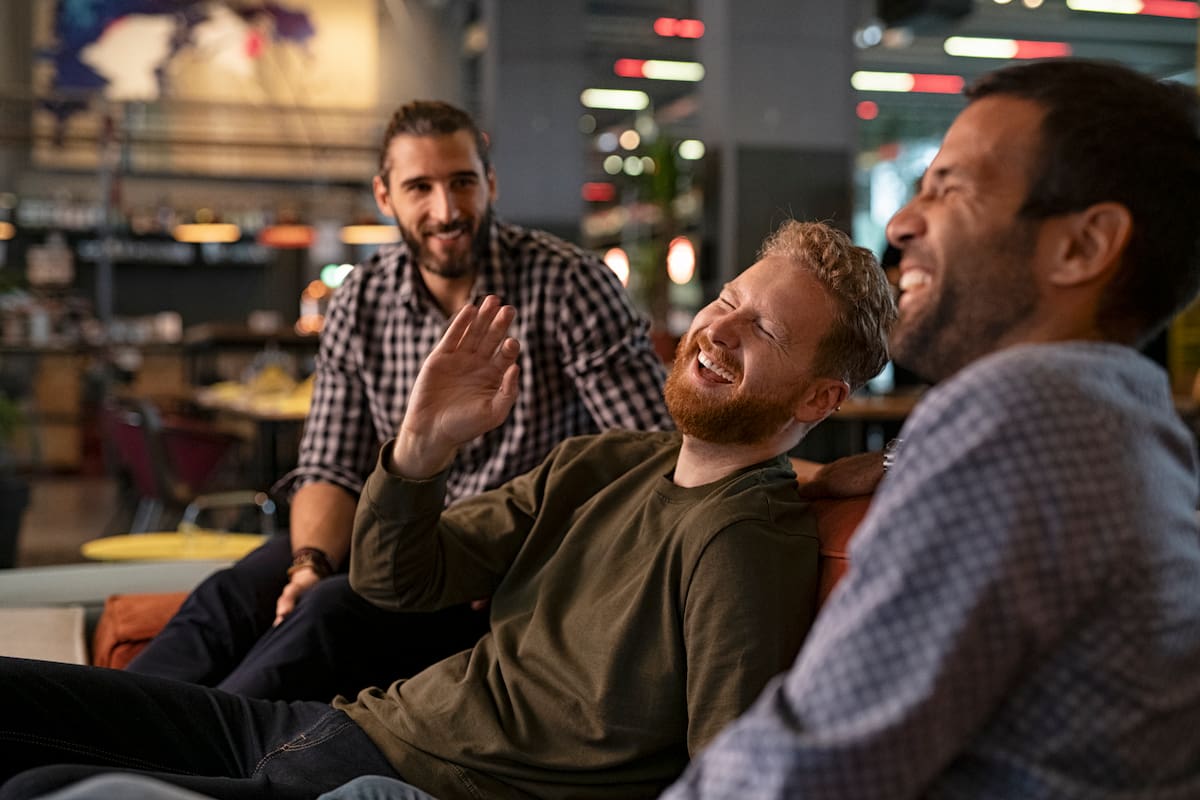 Group of three best friends laughing and enjoying the evening at pub. Happy young men enjoying late night staying together at bar. Cheerful guys sitting on couch and having fun while relaxing.