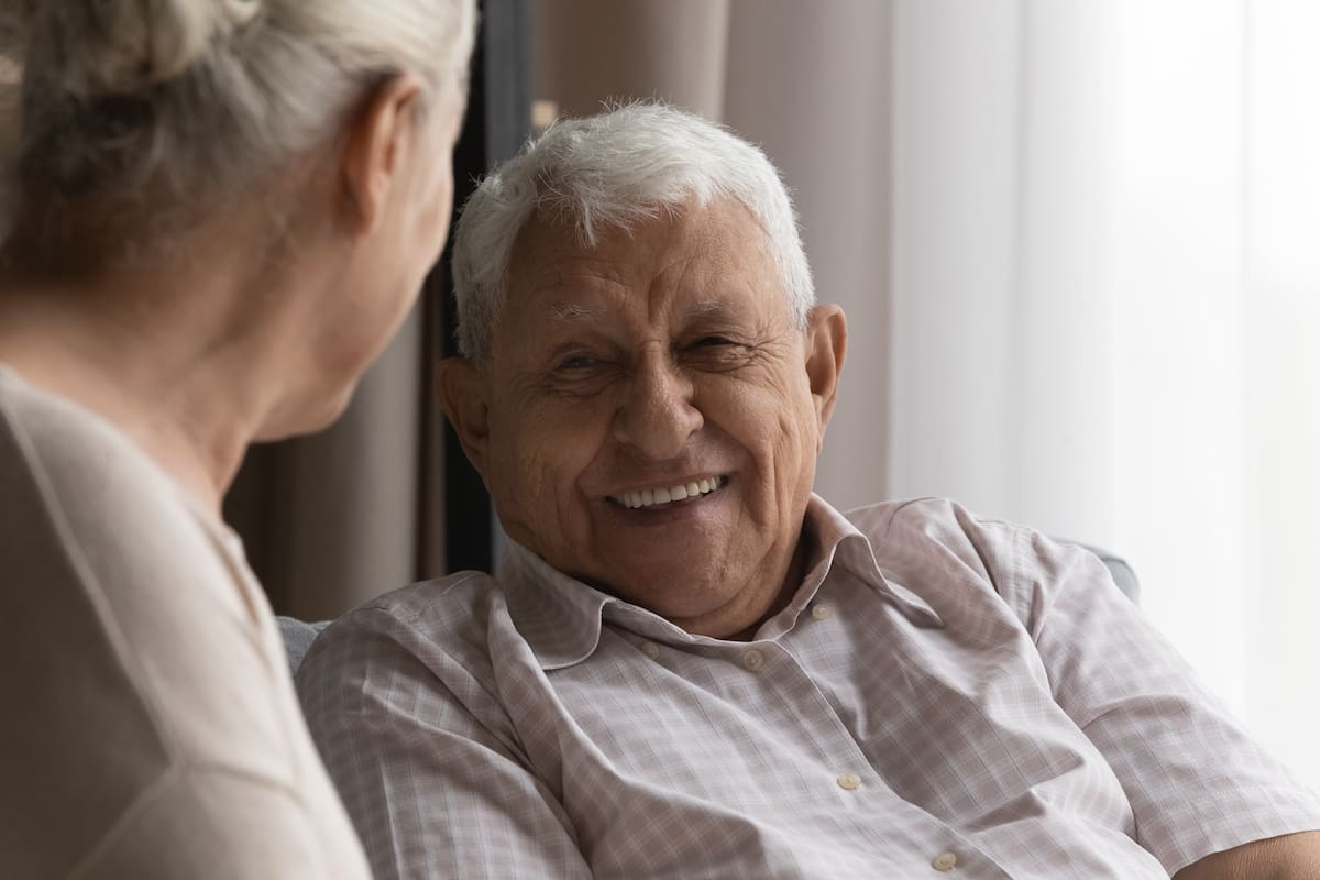 Happy elderly 80s man talking to wife, female carer at home, resting on couch, speaking to woman with toothy smile. Later life, old age, elderly care, marriage concept