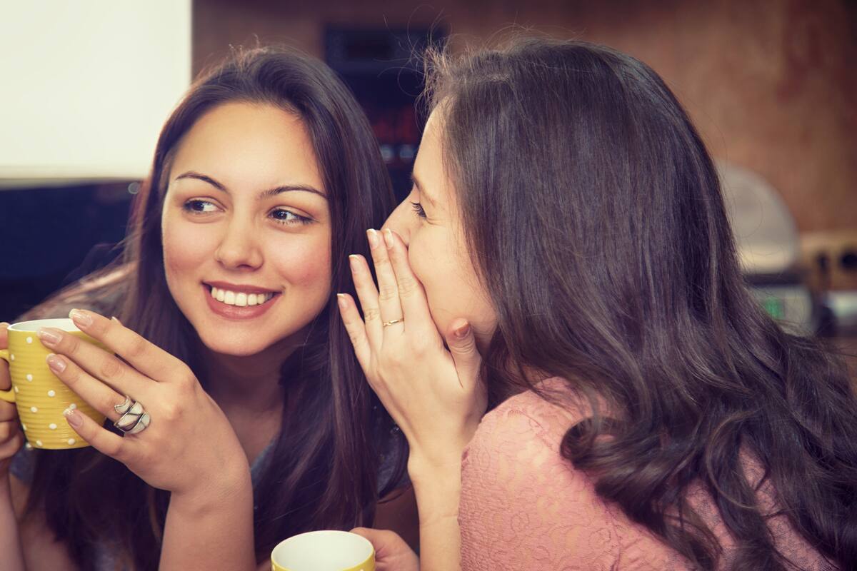 Happy women whispering and drinking coffee. Smiling girls telling secrets and gossiping.
