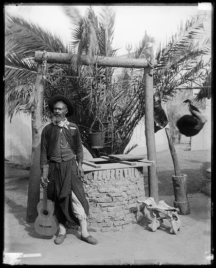 Hombre con atuendo de gaucho y guitarra posa junto a un aljibe. 1902