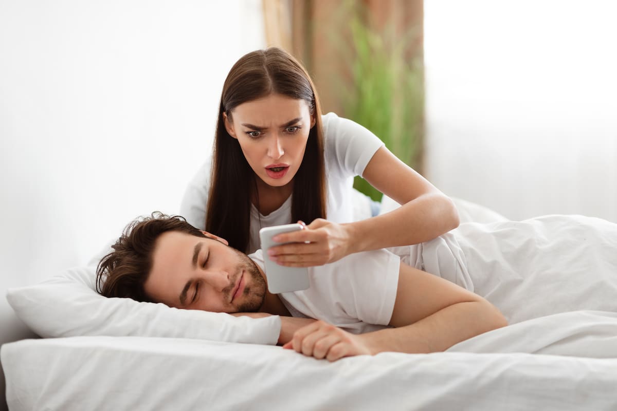 Infidelity. Shocked Wife Reading Message From Lover On Phone While Cheating Husband Sleeping In Bedroom At Home. Jealous Woman Checking Boyfriend's Cellphone Chats. Affair, Relationship Issues