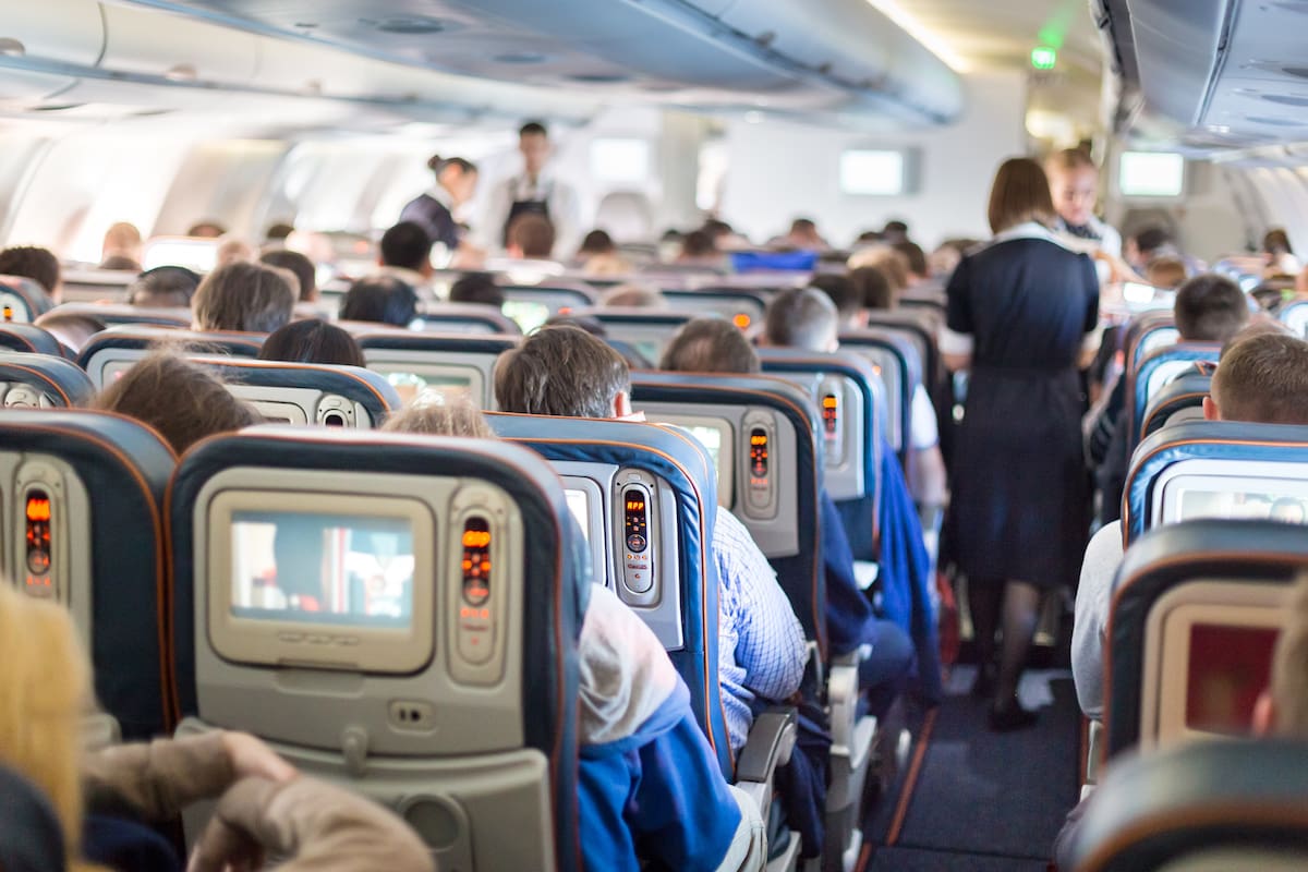 Interior of large passengers airplane with people on seats and stewardess in uniform walking the aisle.