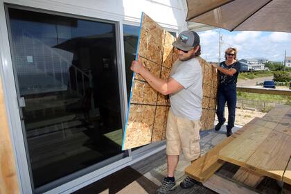 James Masog, al centro, y Gary Tavares, llevan una tabla de conglomerado para proteger las puertas de vidrio de la casa de un cliente en Charlestown, Rhode Island, antes de la llegada del huracán Henri, e sábado, 21 de agosto del 2021. (AP Foto/Stew Milne)